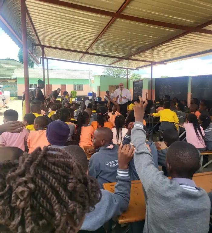Pupils raising their hands at an outdoor assembly, with Welcome to Quick Step School Limited written on the blackboards